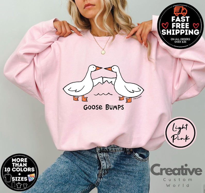 Puede incluir: Sudadera rosa claro con un gr&aacute;fico de dos gansos bes&aacute;ndose y el texto "Goose Bumps".