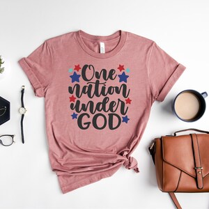 Op de afbeelding: Een roze T-shirt met de tekst "One nation under GOD" in zwarte letters omringd door rode en blauwe sterren.