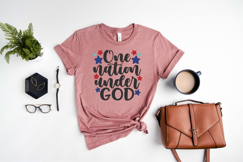 Op de afbeelding: Een roze T-shirt met de tekst "One nation under GOD" in zwarte letters omringd door rode en blauwe sterren.