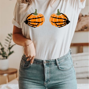 May include: White t-shirt with a graphic of two orange pumpkins with black skeletal hands holding them.