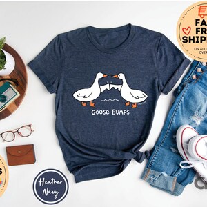 以下が含まれることがあります： ヘザーネイビーブルーのTシャツに、ぶつかり合う2羽のガチョウのグラフィックがプリントされています。ガチョウの下には「Goose Bumps」というテキストがプリントされています。