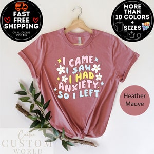 May include: Heather mauve t-shirt with the text "I CAME I SAW I HAD ANXIETY SO I LEFT" in a colorful, playful font. The design includes flower and star graphics. The shirt is displayed on a wooden hanger.