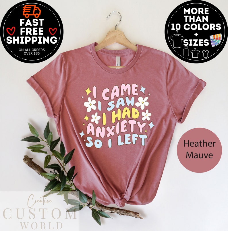 May include: Heather mauve t-shirt with the text "I CAME I SAW I HAD ANXIETY SO I LEFT" in a colorful, playful font. The design includes flower and star graphics. The shirt is displayed on a wooden hanger.
