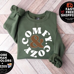 May include: A green sweatshirt with a white circle design that says "Comfy & Cozy" in a retro font. The ampersand is brown.