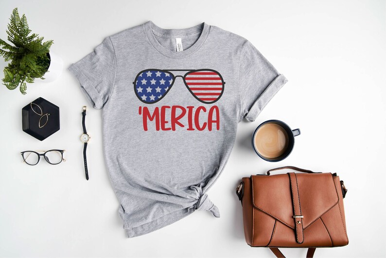 Puede incluir: Camiseta gris con un gr&aacute;fico de gafas de sol con un dise&ntilde;o de bandera estadounidense. El texto 'MERICA est&aacute; impreso debajo de las gafas de sol.