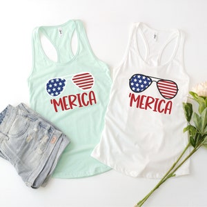 Puede incluir: Dos camisetas sin mangas con el texto 'MERICA' y una imagen de gafas de sol con un dise&ntilde;o de bandera estadounidense. Las camisetas son una blanca y una azul claro.
