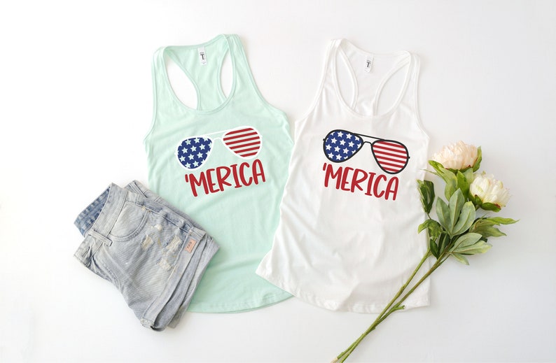 Puede incluir: Dos camisetas sin mangas con el texto 'MERICA' y una imagen de gafas de sol con un dise&ntilde;o de bandera estadounidense. Las camisetas son una blanca y una azul claro.