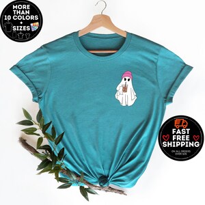 May include: Teal blue t-shirt with a ghost wearing a pink beanie and holding a brown and white cup. The ghost has two black eyes and a small mouth.