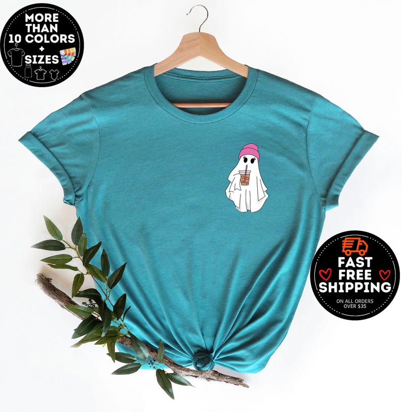May include: Teal blue t-shirt with a ghost wearing a pink beanie and holding a brown and white cup. The ghost has two black eyes and a small mouth.