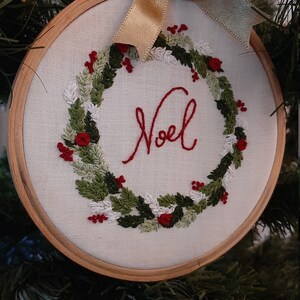 May include: A white embroidery hoop with a green and red wreath and the word "Noel" embroidered in red thread. The hoop is decorated with a gold ribbon bow.