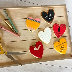May include: Six heart-shaped cookies decorated with school-themed designs. The cookies feature a pencil, a ruler, a piece of paper, a red crayon, a worm, and a chalkboard with the text "Thank You!"