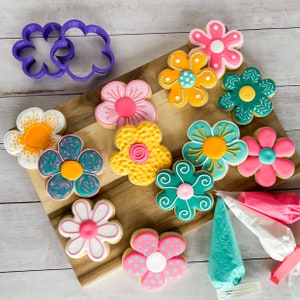 May include: A wooden board with nine decorated sugar cookies in the shape of flowers. The cookies are decorated with various colors, including pink, yellow, green, and blue. There are also three piping bags with frosting in pink, white, and blue.