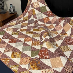 May include: A patchwork quilt featuring a repeating pattern of triangles and squares in shades of rust, mustard yellow, sage green, and cream. The fabric displays various floral, plaid, and geometric prints with visible machine quilting stitches.