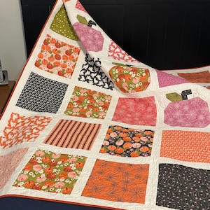 May include: A patchwork quilt featuring a grid of square fabric blocks with various seasonal prints including pumpkins, spiderwebs, ghosts, and stripes in orange, black, pink, and green. The quilt has a white background and an orange border.