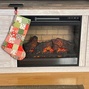 May include: A Christmas stocking featuring a patchwork design of various fabric squares in shades of red, green, white, and gray. The stocking has a green fabric cuff at the top and a small red loop for hanging, displaying floral and geometric patterns.