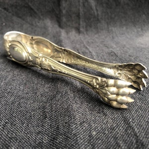 May include: Ornate silver sugar tongs with a detailed floral and scroll design. The tongs have a classic shape with a squeeze-to-open mechanism and claw-like ends. The metal has a polished finish.