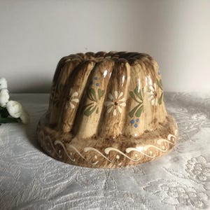May include: A brown ceramic bundt cake pan with a white and green floral design. The pan has a fluted edge and a decorative border.