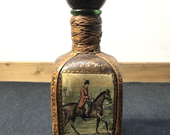 May include: A brown leather-wrapped liquor bottle with a round wooden stopper. The bottle features a vintage illustration of a man riding a horse with a dog running in the foreground.