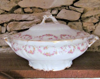 May include: A white porcelain serving dish with a lid, featuring a delicate floral pattern in shades of pink and gold. The dish has a scalloped edge and ornate handles.