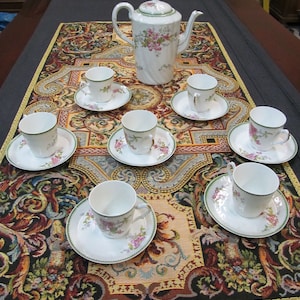 May include: A white porcelain tea set with pink floral accents, including a teapot and six teacups with saucers. The set is arranged on a decorative tapestry tablecloth with a dark background and ornate patterns.
