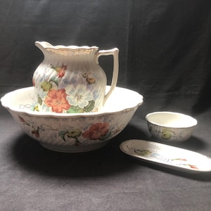 Könnte beinhalten: Ein weißes Porzellan-Set aus Krug, Schüssel und Seifenschale mit einem Blumenmuster in Rosa-, Blau- und Grüntönen. Das Set hat einen gewellten Rand und ein zartes Blumenmuster.
