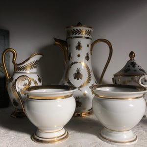 Op de afbeelding: Een set witte porseleinen theekopjes en een theepot met gouden accenten en bijendesigns. De theepot heeft een deksel en een handvat. De kopjes hebben een ronde vorm en een handvat. De set staat op een wit oppervlak.