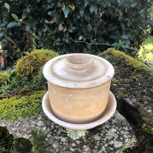 Könnte beinhalten: Eine antike, cremefarbene Butterdose aus Keramik mit Deckel und separatem Sockel. Die Dose hat ein schlichtes, elegantes Design mit einem erhöhten Rand und einem dezenten Muster.