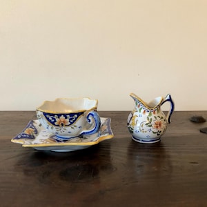 May include: A ceramic teacup and saucer set with a matching creamer. The teacup and saucer are white with blue and yellow floral designs. The creamer has a blue handle and a similar floral pattern.