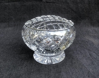 May include: A clear, cut-glass flower frog with a diamond pattern and a metal grid lid. The round bowl sits on a circular base. This decorative piece is designed for arranging flowers.