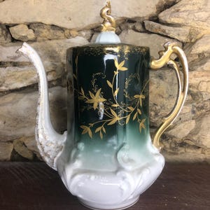 Puede incluir: Una tetera de porcelana blanca y verde con detalles dorados y un diseño floral. La tetera tiene una tapa con un pomo dorado y un asa con un detalle dorado.