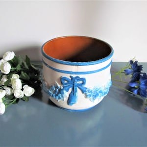 Könnte beinhalten: Weißer Keramiktopf mit einem blauen Schleifendesign und blauen floralen Akzenten. Der Topf hat ein braunes Terrakotta-Innere.
