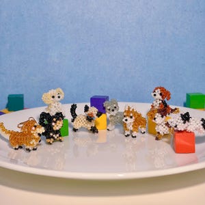 May include: A collection of beaded dog figurines in various breeds and colors, including tan, black, white, and gray. The dogs are displayed on a white plate with colorful blocks in the background.