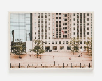 May include: A photograph of a large building with a glass facade and a brick facade. The building has many windows and a flag flying from the top. There are trees in front of the building and people walking on the pavement.
