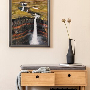 May include: A wooden bench with two drawers and a gray cushion. A gray throw blanket is draped over the bench. A black vase with dried flowers sits on top of a stack of books on the bench. A gray woven basket sits on the floor in front of the bench. A framed photograph of a waterfall in a landscape setting hangs on the wall behind the bench.