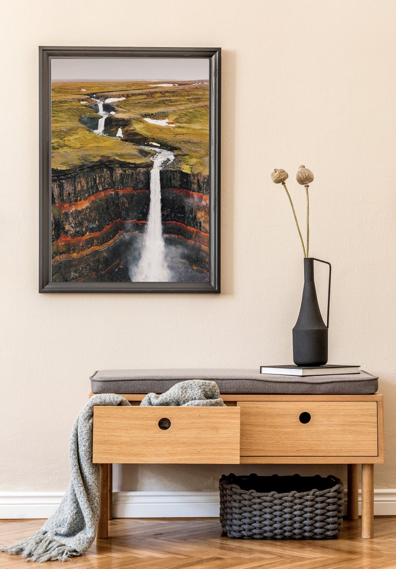 May include: A wooden bench with two drawers and a gray cushion. A gray throw blanket is draped over the bench. A black vase with dried flowers sits on top of a stack of books on the bench. A gray woven basket sits on the floor in front of the bench. A framed photograph of a waterfall in a landscape setting hangs on the wall behind the bench.