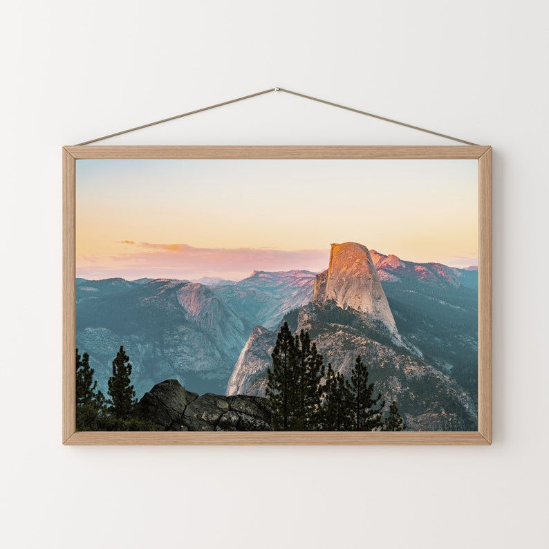 May include: A framed photograph of a mountain range with a large rock formation in the center. The sky is a soft pink and orange, and the mountains are a deep blue and green. The photo is framed in a light wood frame.
