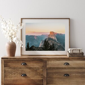 May include: A framed photograph of Half Dome in Yosemite National Park, California. The photo shows the iconic rock formation at sunset, with a soft pink and orange glow in the sky. The photo is framed in a simple, modern frame and is displayed on a wooden dresser.