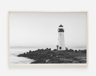 May include: A black and white photograph of a lighthouse on a rocky shore. The lighthouse is tall and white with a black roof and a black lantern. The rocks are dark and jagged, and the water is choppy.