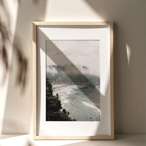 Puede incluir: Una fotograf&iacute;a enmarcada de una escena de bosque brumoso con una playa y el oc&eacute;ano en primer plano. La foto est&aacute; en un marco de madera clara y est&aacute; colgada en una pared blanca.