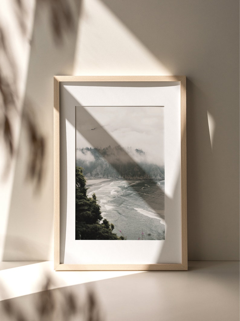 Puede incluir: Una fotograf&iacute;a enmarcada de una escena de bosque brumoso con una playa y el oc&eacute;ano en primer plano. La foto est&aacute; en un marco de madera clara y est&aacute; colgada en una pared blanca.