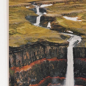 May include: A cascading waterfall flows over a rocky cliff face, with green grass and snow patches in the background.