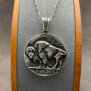 May include: A silver pendant necklace featuring a detailed depiction of a buffalo on a coin. The coin is inscribed with "UNITED STATES OF AMERICA", "E PLURIBUS UNUM", and "FIVE CENTS". The pendant hangs from a delicate silver chain.