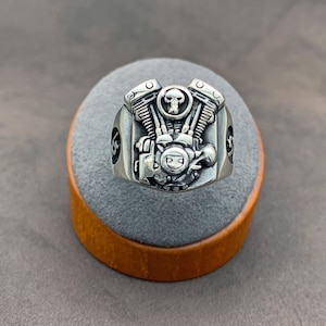May include: A silver ring with a detailed motorcycle engine design. The engine has a skull in the center and is surrounded by intricate details. The ring is on a gray and brown display stand.