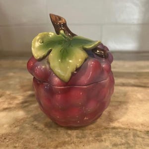 May include: A ceramic jar shaped like a cluster of grapes. The jar is primarily red with purple accents, a green leaf, and a brown stem. The lid is designed to look like the top of the grape cluster.
