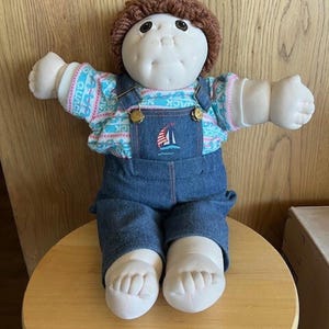 May include: A vintage doll with a soft, vinyl face and brown yarn hair. The doll wears a blue and white patterned shirt under denim dungarees with a sailboat emblem. The doll is seated on a wooden stool.
