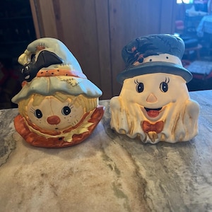 May include: Two ceramic Halloween figurines. One is a scarecrow with a blue hat and a black bat. The other is a ghost wearing a top hat and a bow tie. Both have smiling faces and are painted in bright colors.
