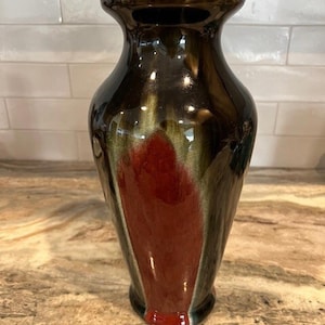 Puede incluir: Un jarrón de cerámica alto y brillante con un borde acampanado. El jarrón presenta un exterior marrón oscuro con un diseño central rojo y verde. El jarrón es adecuado para exhibir flores o como elemento decorativo.