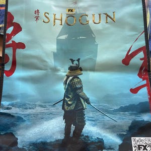 Puede incluir: Bolsa tote de color azul claro con un guerrero samurái con una espada en el océano. La palabra "SHOGUN" está impresa en dorado, con caracteres japoneses y el logo de FX. Un castillo se ve en el fondo.