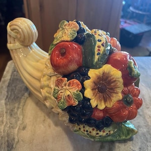 Puede incluir: Una cornucopia de cerámica rebosante de coloridas y detalladas frutas y verduras. La bocina es de color amarillo cremoso y el arreglo incluye manzanas rojas, uvas, maíz y girasoles. Una pieza decorativa para el hogar.