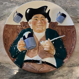 Puede incluir: Plato decorativo de cerámica que representa a un hombre jovial con sombrero tricornio y abrigo verde, sosteniendo una jarra azul. El plato tiene un fondo crema con dos pequeñas jarras azules y detalles marrones. Objeto de colección vintage.